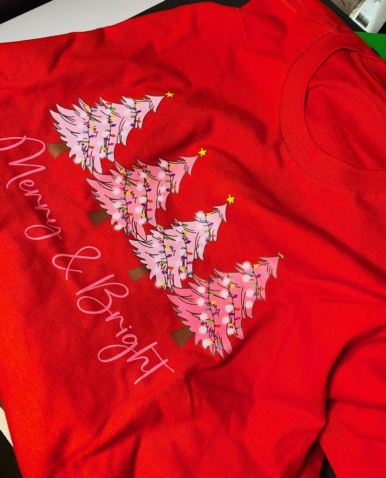 The image shows a festive red t-shirt with a unique Christmas tree design made of pink flamingos, each topped with a yellow star. The phrase “Merry & Bright” is written in cursive white text, adding to the holiday spirit of the image. It’s a creative take on traditional Christmas t-shirts. Gallery Display